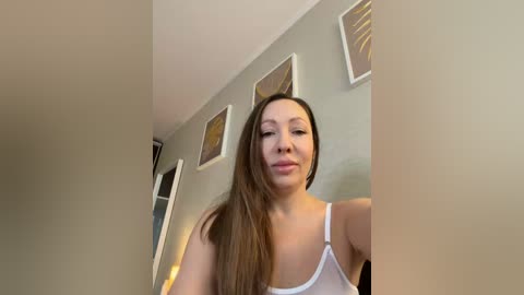 Video of a young Asian woman with long straight brown hair, wearing a white tank top, standing in a dimly lit hallway with beige walls and abstract art.