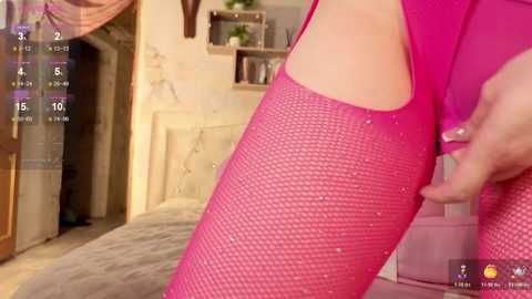 Video of a woman wearing bright pink fishnet stockings, standing in a bedroom with a bed, a fireplace, and a shelf in the background.
