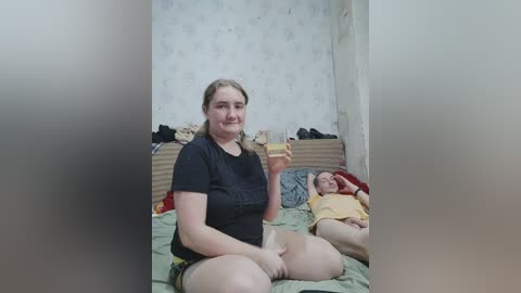 Video of a young woman with light skin and brown hair, sitting on a bed in a cluttered room. She wears a black T-shirt and shorts, holding a drink. A baby lies beside her in a yellow onesie.