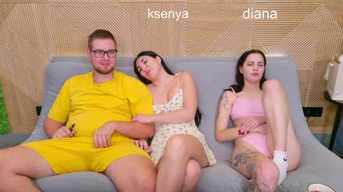 Video of a young man and two young women, one in a yellow t-shirt, one in a floral dress, and one in pink lingerie, sitting on a modern grey couch against a wooden backdrop.
