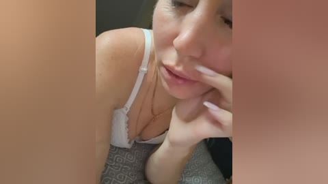 A video of a light-skinned woman with a slender physique, wearing a white lace bra, lying on a patterned gray fabric. She gently touches her lips with her fingers, her expression soft and contemplative.