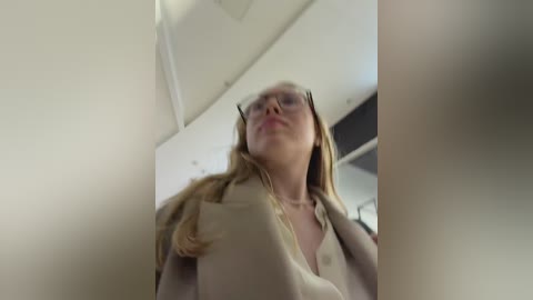 Video of a young Caucasian woman with light skin, blonde hair, wearing glasses and a beige top, looking up in a dimly lit room with white walls and a ceiling.