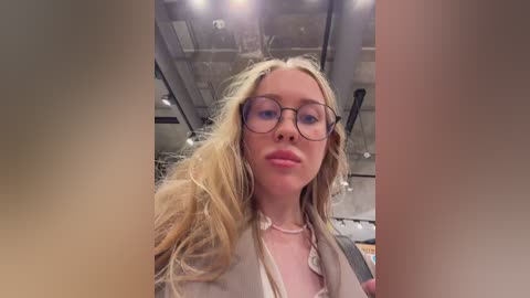 A video of a young, fair-skinned woman with long, wavy blonde hair, wearing round glasses and a beige blazer, taken indoors under fluorescent lighting.