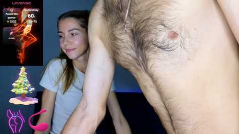 A video shows a young woman with light skin and brown hair, wearing a white shirt, looking at a man's hairy chest. Background includes a Christmas tree and a pink flamingo.