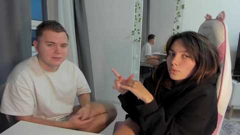 Video of a white man and woman seated in a modern, minimalist living room. The man wears a white t-shirt, and the woman, with long brown hair, a black hoodie. They gesture and talk, with a mirror reflecting another person in the background.