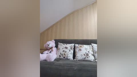 Video of a cozy living room with a gray fabric sofa, adorned with light floral cushions, and a large, fluffy pink teddy bear placed on the left side. The background features a slanted wooden wall paneling in a light beige color.