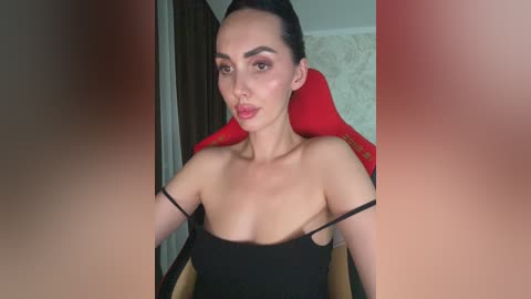Video of a woman with light skin and dark hair, wearing a black strapless dress, sitting in a red gaming chair in a room with beige wallpaper.