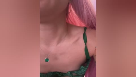 A video of a fair-skinned woman with long pink hair, wearing a green lace bra with a dangling emerald pendant necklace, partially visible. The image is cropped close, focusing on her bare shoulders and chest.