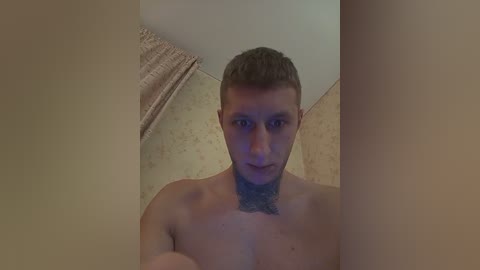 A video of a shirtless, muscular man with short brown hair, a light beard, and a tattoo on his chest. The background features a textured beige wall and a partially visible window with a beige curtain. The image is dimly lit.