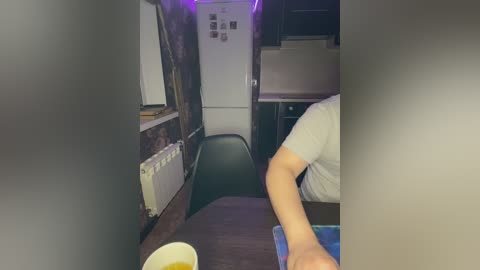 A video of a narrow, dimly-lit, cramped RV kitchen with worn, brown walls. A person in a white shirt sits at a small table, holding a glass of beer.