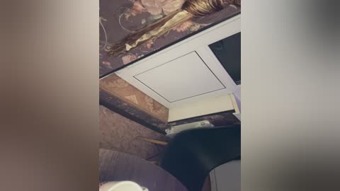 Video of a woman's head and upper torso, partially obscured by a beige curtain. She has light skin, short hair, and is wearing a black top. The background features a floral-patterned wall and a white ceiling panel.