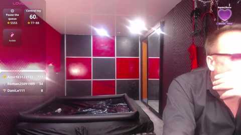 A video of a dimly lit, black-and-red tiled room with a black coffin in the foreground. A man with glasses and a dark shirt stands in the right background, watching a live stream.