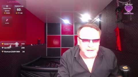 A video of a white man with short brown hair, wearing dark sunglasses and a black shirt, live-streaming on a gaming platform. The background features a black and red gaming setup, with a pool table and a monitor displaying game statistics.
