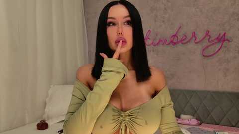 Video of Kim Kardashian, a fair-skinned woman with long black hair, wearing an off-shoulder green top, making a finger-to-mouth gesture, in a cozy bedroom with pink \"Kimberly\" graffiti on the wall.