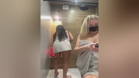 A video captures a blonde woman with a black face mask, wrapped in a towel, walking away in a dimly lit, wooden-paneled sauna.