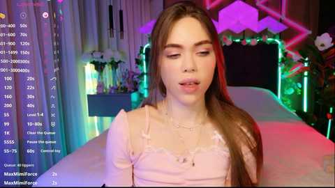 Video of a young woman with fair skin, long brown hair, and light pink lingerie, sitting on a bed in a brightly lit room with neon lights and floral decorations.