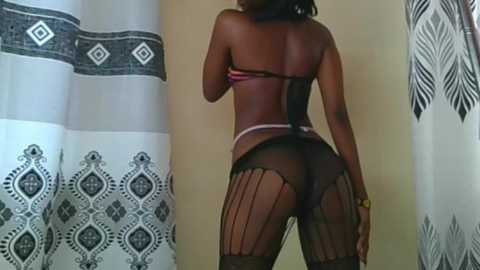 Video of a black woman with dark skin, wearing a sheer black bodysuit, standing in a bathroom with patterned shower curtains.