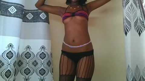 Video of a slim, dark-skinned woman with medium-length black hair, wearing a striped bikini top, black fishnet stockings, and matching panties, standing in a room with patterned curtains and a beige wall.