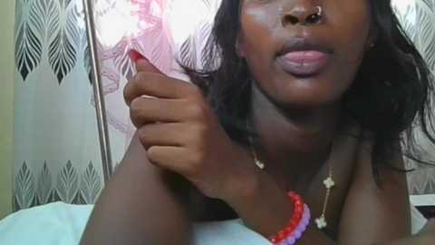 Video of a dark-skinned woman with wavy black hair, wearing a nose ring, red bracelet, and necklace, holding a lighter, against a patterned white and black background.