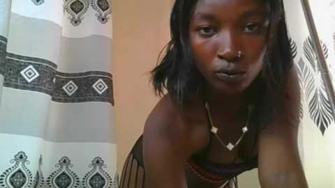 Video of a dark-skinned woman with shoulder-length black hair, wearing a black dress with a beaded necklace. Background features a patterned curtain and a beige wall.