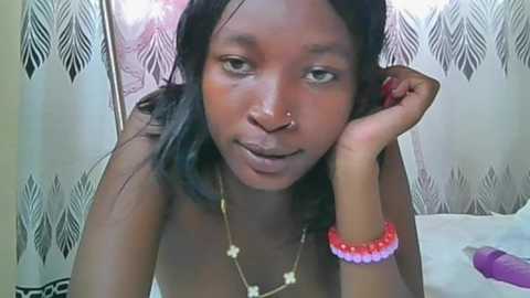 Video of a young black woman with medium skin tone, wearing a nose ring, sitting on a bed with a white sheet, against a background of patterned curtains.