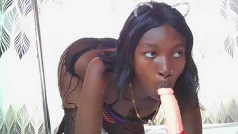 Video of a dark-skinned woman with long black hair, wearing black lingerie, performing oral sex on a large, pink dildo. Background features a patterned, light-colored curtain.