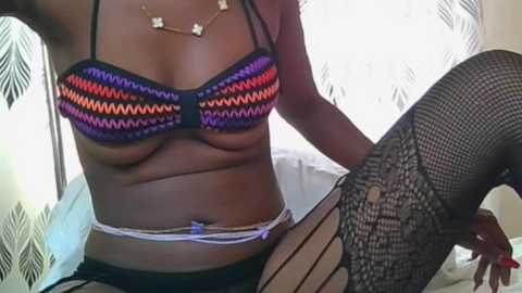 Video of a dark-skinned woman in a colorful, patterned bikini top and black lace fishnet stockings, sitting on a bed with white sheets, against a patterned curtain background.