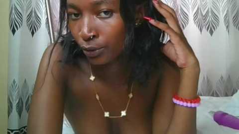 Video of a topless African-American woman with dark skin and long black hair, wearing a gold necklace and colorful bracelets, sitting on a bed with white sheets and a black-and-white feather-patterned curtain background.