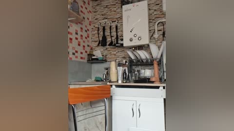 Video of a cozy kitchen with a white sink, wooden chair, and rustic stone backsplash. Stainless steel utensils hang on a wall rack. A white oven and microwave are integrated into the counter.