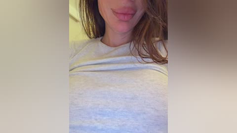 A close-up video of a Caucasian woman with fair skin, brown hair, and a light grey shirt. She is smiling with her tongue slightly out. The background is blurred and beige-colored.