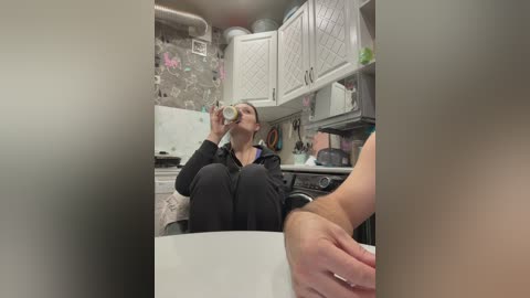 Video of a woman in a kitchen, wearing a black hoodie, drinking coffee, white cabinets, grey tiled backsplash, and a hand resting on a white countertop.