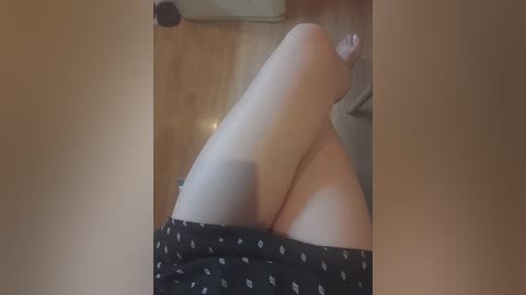 Video of a woman's bare legs in black pajama shorts, sitting on a wooden floor, with a blurred background.