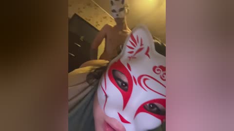 A video of a person wearing a white mask with red patterns and a yellow shirt, taken from an angled, close-up perspective.