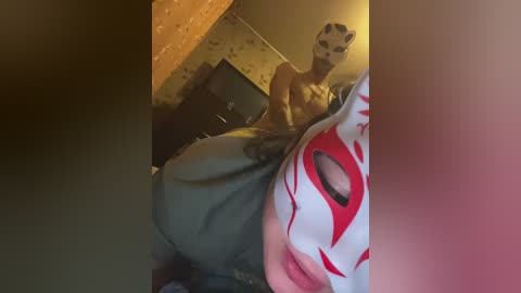 Video: A person in a white mask with red markings and a gray costume, standing in a dimly lit room with patterned wallpaper.