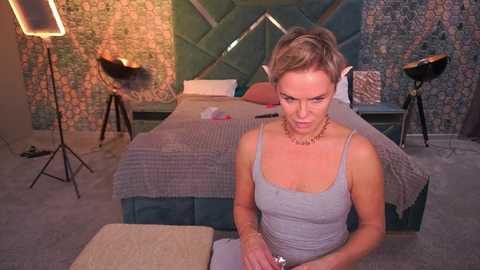Video of a middle-aged woman with short blonde hair in a gray tank top, seated at a bed with floral wallpaper and a green quilt, surrounded by studio lights and a tripod.
