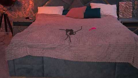 A video of a cozy, rustic bedroom featuring a large bed with a knitted pink blanket embroidered with a spider, teal pillows, and a brick wall backdrop.