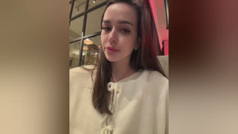 Video of a young woman with long brown hair, fair skin, and a neutral expression, wearing a white robe, standing in a dimly lit room with a red wall and a window with reflective glass.