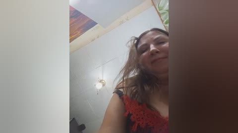 Video of a middle-aged woman with medium-length brown hair, wearing a red lace top, smiling slightly, standing in a dimly lit room with a white wall, a wooden ceiling, and a hanging light fixture.