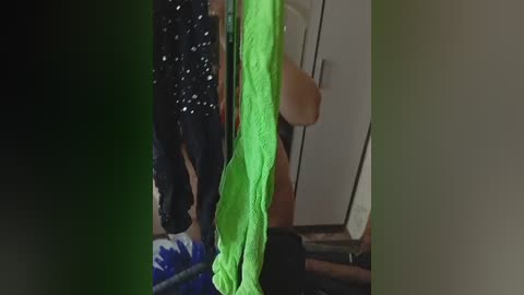 Video of a woman with long, dark hair wearing a bright green towel, standing in a dimly lit room with a black jacket and a blue patterned skirt visible in the background.