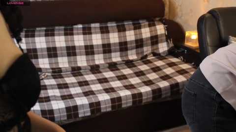 Video of a cozy, vintage-style sofa with a brown and white checkered fabric cover. The sofa sits against a beige wallpapered wall, and a person in a white shirt is partially visible in the background.