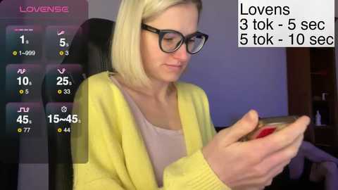 Video of a blonde woman with glasses, wearing a yellow cardigan, playing a game on a smartphone. Background shows a purple and black gaming chair, a TV screen displaying \"Love\" and game stats.