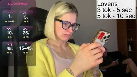 Video of a blonde woman with glasses, wearing a yellow cardigan, sitting on a black chair, scrolling on a smartphone. Text overlay: \"Lovenese\" and \"Lovens 3 tok-5 sec.\
