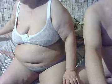 A video shows a plus-sized woman in a white bra and panties, sitting on a bed with a patterned sheet. Her skin is light, and she has a soft, rounded belly. The background features a tiled wall.
