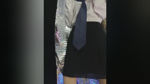 Video of a woman wearing a white blouse, black skirt, and blue tie, standing in a room with foil-covered objects on a table.