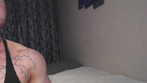 A video showing a person with a tattoo on their left shoulder, partially visible, standing beside a bed with white sheets, against a dark, patterned curtain and a beige wall with framed pictures.