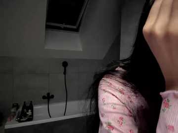 Video of a person with long black hair, wearing a pink floral blouse, standing in a dimly lit bathroom with a tiled ceiling, white bathtub, and a showerhead on the wall.