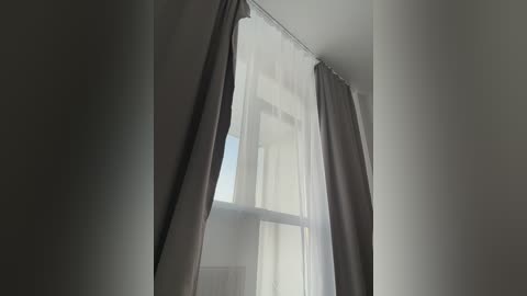 Video of a minimalist room with a large window adorned by sheer white curtains and grey curtains on either side, allowing soft daylight to filter through, casting gentle shadows on the plain white walls.