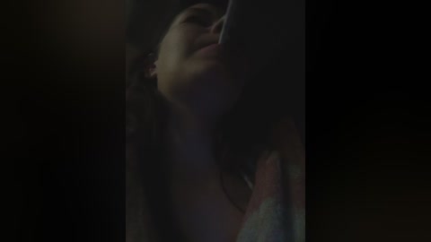 A dimly lit, close-up video of a woman with long dark hair and closed eyes, leaning against a dark background, possibly indoors, creating a moody, intimate atmosphere.