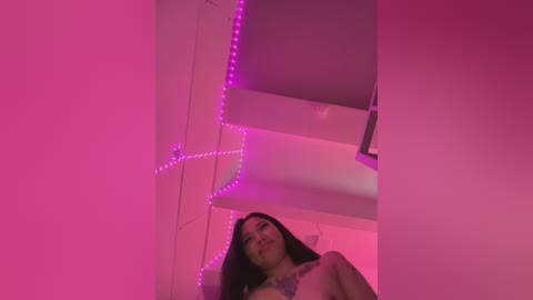 A video of a nude woman with long dark hair, under a pink LED light, lying on a bed in a dimly lit room.