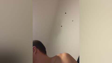 Video of a shirtless man with short dark hair, facing away, in a dimly lit room with a beige wall and bullet holes.